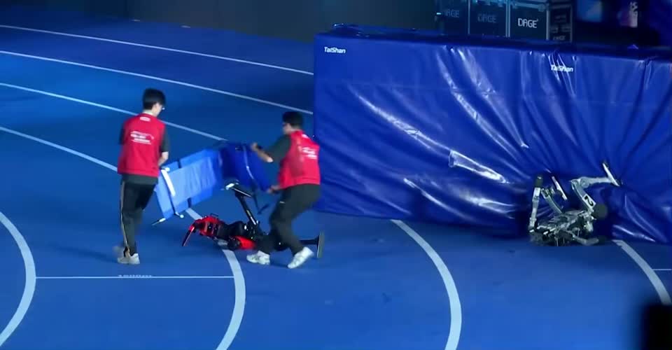 Frame aos 26 s: pós-chegada, um robô vermelho na pista coberto por um colchão pelo staff e um robô prateado dentro do colchão da parede; nenhuma chama à vista.
